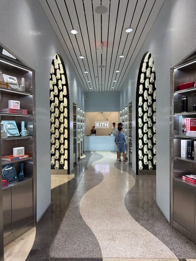 The image depicts the interior of a Kith store in Miami Beach, Florida, United States of America. The scene is a long, narrow corridor with high ceilings featuring a slatted design and recessed lighting. The walls are a light, textured gray, and the floor is a patterned terrazzo with wavy, contrasting bands of dark and light gray.
On either side of the corridor, floor-to-ceiling display units are visible. The left side features stainless steel shelving stocked with various items, including books and what appear to be lifestyle products. The right side also has stainless steel shelving, showcasing books and other merchandise.
Dominating the view are two large, arched niches on the walls. Each niche contains a vertical arrangement of numerous white sneakers, illuminated from within, serving as a prominent design element.
In the background, a counter area is visible with a Kith logo displayed prominently. Two individuals, likely employees, are behind the counter. Two young boys, dressed in blue, are standing near the center of the corridor, facing towards the counter, suggesting they are customers. One of the boys is looking towards the counter, while the other is looking downwards. The overall atmosphere is clean, modern, and retail-focused.