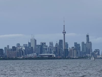 The media file displays a panoramic view of the Toronto skyline on an overcast day. The scene is dominated by a dense collection of skyscrapers of varying heights and architectural styles, creating a striking urban silhouette against a muted, cloudy sky. Prominent structures include the iconic CN Tower, which stands tall and slender at the center of the skyline, and other modern high-rises.
In the foreground, a body of water, likely Lake Ontario, stretches across the bottom of the frame. The water's surface is rippled, suggesting a gentle breeze. A small white sailboat with black trim is visible on the water to the right of the frame, adding a touch of activity to the otherwise still scene.
Along the waterfront, a strip of lush green trees forms a natural boundary between the city and the lake. Behind the trees, parts of the cityscape are visible, including what appears to be a stadium with a distinctive white roof structure and a wind turbine. The overall impression is one of a grand, modern city viewed from a distance across the water. The overcast weather contributes to a somewhat subdued color palette and a sense of calm, perhaps a quiet afternoon. There is no visible text in the image.