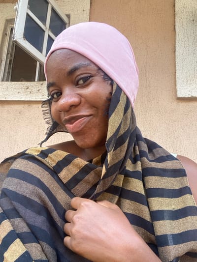 A woman with dark skin and a pink head covering looks at the camera. She wears a black and gold striped shawl or shirt. Part of a window is visible in the background, along with a beige or light brown exterior wall. The woman's head is tilted to the side.