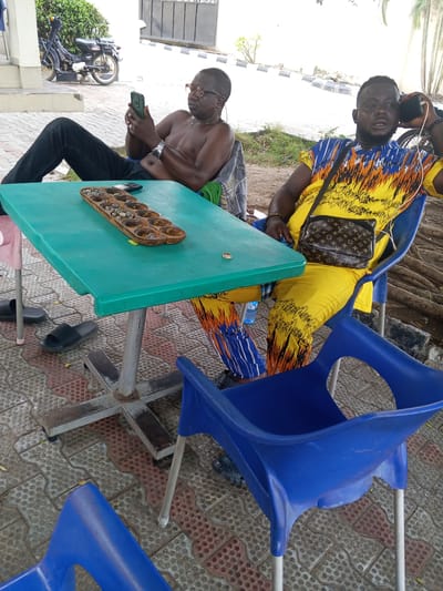 The image depicts two men seated at a green table on a patio in Lokoja, Nigeria. A wooden mancala board with game pieces sits on the table. One man is shirtless, wearing glasses and dark pants, and is looking at a smartphone. The other man wears a yellow and blue patterned outfit and has a Louis Vuitton crossbody bag. He is holding a phone to his ear. Blue plastic chairs surround the table. In the background, there is a parked motorcycle, a building with a metal gate, and some greenery. The ground is paved with patterned interlocking bricks.