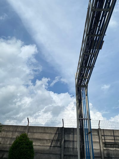 In Gambut Barat, Indonesia, a concrete wall with barbed wire atop it runs horizontally across the lower half of the frame, casting shadows. To the left, a small, dark green bush is situated in front of the wall. Dominating the right side and extending upwards is a metal structure supporting numerous black cables, some of which are encased in blue conduits. The cables ascend at an angle against a cloudy sky with patches of blue.