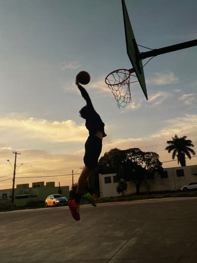 A silhouette of a young person is captured mid-air, jumping to dunk a basketball. The person is wearing dark clothing and brightly colored sneakers, one red and one green, suggesting dynamic movement. The basketball is held high in their right hand, aimed towards the hoop. The basketball hoop, with its net, is visible against a hazy sky filled with pale orange and blue clouds, indicative of either sunrise or sunset. In the background, several buildings with windows are silhouetted against the sky. A street light and power lines are also visible, hinting at an urban or suburban setting. A palm tree stands tall on the right side of the frame. A silver car is parked on the street in front of one of the buildings, and a yellow car is driving on the road. The foreground shows a paved surface, likely a basketball court. The overall scene is set outdoors during twilight.