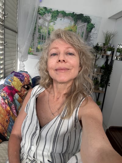 A selfie shows a middle-aged woman with blonde, wavy hair, wearing a striped top and a necklace, indoors. Behind her are various items, including a painting of a garden scene, a shelf with plants, and a quilted blanket. The location is purportedly in Ramat Gan, Israel, though there is no visual confirmation of this location.