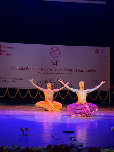 Two male dancers are performing on a stage at the Tagore Theatre in Chandigarh, India, at the 54th All India Bhaskar Rao Nritya & Sangeet Sammelan. The event is being held from 26th to 31st March, 2025, and is supported by the Ministry of Culture, Government of India. Both dancers are wearing traditional Indian attire, with one in an orange dhoti and the other in a purple dhoti. They are both in a seated position, with their arms outstretched in a dance pose. The stage is lit with blue lights and there are garlands and a black cable on the floor in front of the dancers.  The backdrop is a white banner with the event details and logo.  The coordinates provided are for the Tagore Theatre in Chandigarh, India.