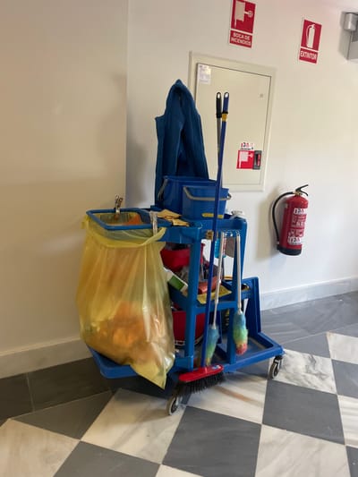 A blue cleaning cart is positioned in a hallway in Segovia, Spain. The cart is loaded with cleaning supplies including a yellow plastic garbage bag filled with debris, a blue bucket, and a blue mop with a white and rainbow-colored brush. The cart is situated in a hallway with white walls, a black and white checkered floor, and two red fire extinguishers mounted on the wall. The image was captured at latitude 40.953, longitude -4.12. A blue jacket is hanging from the wall, partially obscuring the fire extinguishers. The scene is clean and organized.
