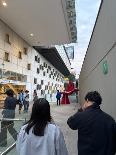 The image, taken in Mexico City, Mexico, shows a pedestrian walkway between two buildings under an overcast sky. A red, stylized sculpture of two hands is positioned along the walkway. Several people are present: one in a blue jacket, two taking photos near the entrance of a building, and two more partially visible in the foreground. The architecture is modern, with a white building featuring rows of small windows and glass paneling. A large advertisement is visible atop one of the buildings.
