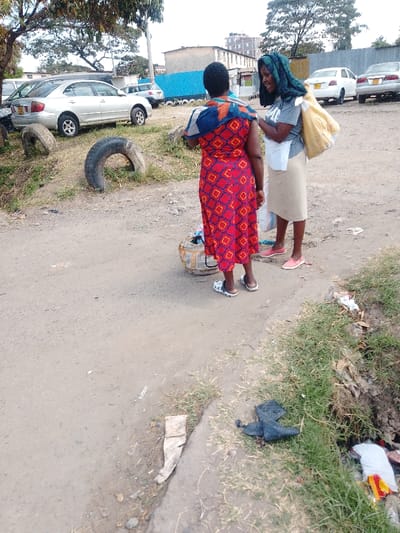 The scene depicts an unpaved urban or semi-urban environment in Nairobi, Kenya, during daylight hours. Two adult females are conversing on a dirt path, positioned roughly center-right.

The woman on the left, with her back mostly to the camera, is wearing a long, red dress with a repeating geometric pattern of red rectangles outlined in yellow with blue inner squares. A multi-colored patterned cloth, featuring shades of orange, blue, and brown, is draped over her shoulders. She wears white and black open-toed sandals. At her feet, a small, cylindrical object, possibly a bucket or container, wrapped in a light-colored material, rests on the ground.

The woman on the right faces the first woman, smiling. She wears a dark head covering, a grey t-shirt, and a light-colored, knee-length skirt (beige or khaki). Pink flat shoes are visible on her feet. A large yellow sack or bag is slung over her left shoulder, and she holds a white plastic bag in her left hand.

The ground is composed of light brown soil/dirt, with a grassy verge running along the right side. This verge contains scattered litter, including a dark, folded object and colorful plastic remnants. In the mid-ground to the left, several discarded black car tires are present, some upright, others lying on their side.

Behind the individuals, an unpaved area serves as an informal parking lot. Multiple vehicles are visible, including a silver Toyota Premio or Allion sedan parked prominently to the left. Further back, additional light-colored sedans are partially obscured. A long, light blue corrugated metal fence extends horizontally across the background, separating the parking area from multi-story buildings behind it. These buildings appear to be a mix of residential and commercial structures, some exhibiting signs of wear. Trees with green foliage are scattered throughout the background and upper left quadrant of the image. The sky above is bright and shows some cloud cover.