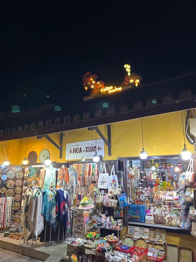 This is an exterior nighttime photograph of a shop in Hội An, Vietnam, which is likely a souvenir or textile shop. The shop features a yellow facade with a dark brown roofline adorned with decorative elements and lights. A sign above the entrance reads "HÒA-XUÂN" in white lettering on a dark background. The shop is packed with various items for sale, including textiles like scarves and clothing draped over mannequins, woven baskets and bags, decorative plates, and an assortment of small trinkets and stuffed animals. Several string lights and decorative lanterns are visible, illuminating the merchandise and the storefront. The overall impression is one of a vibrant and busy night market scene.