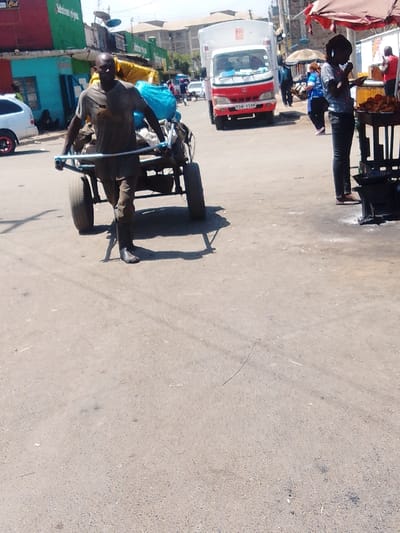 The image depicts a bustling urban street scene in Nairobi, Kenya, under bright daylight. In the foreground, slightly left of center, a dark-skinned adult male with a bald head, wearing a dirty brown t-shirt, dark trousers, and dark rubber boots, actively pulls a heavy, two-wheeled handcart. The handcart is constructed from metal, possibly improvised, and is overloaded with bundled items, including a prominent blue tarp or bag. The man grips a light-colored horizontal bar serving as the handle.

To the right, a lean individual, seen from the side, with dark hair and wearing dark trousers and a light-colored patterned top, stands next to a street vendor's stall. This stall is dark-colored, possibly metal, with a wide, shallow pan containing what appears to be fried food items. A portion of a reddish-brown umbrella providing shade for the stall is visible above. Ash or residue is present on the ground around the base of the stall, indicative of a charcoal burner.

In the midground, a red and white box truck (license plate KBN 958M) is positioned on the asphalt road, facing away from the viewer. To the far left, a white sedan with a red accent on its rear wheel is parked. Multiple individuals are visible in the background, including a person in a blue top behind the truck and another in a red top near the vendor stall.

The left side of the street features a row of single-story commercial buildings or shops with vibrant facades: green, blue, and red. A satellite dish is mounted on the green building. In the background, multi-story buildings, likely residential or mixed-use, complete the urban landscape. The road surface is grey asphalt, appearing worn in areas. Strong shadows are cast by the subjects, indicating direct sunlight from a high angle.