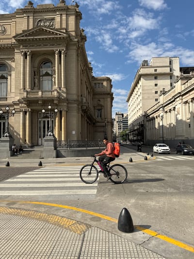 This vertical image captures a bustling street scene in Buenos Aires, Argentina, under a bright, partly cloudy blue sky. The strong sunlight casts distinct shadows, suggesting it's either mid-morning or afternoon.
The primary subject is a person on a black bicycle, standing on a wide, striped pedestrian crossing in the center-right of the frame. They are wearing a peach or light orange hoodie and a prominent bright orange backpack. The bicycle frame clearly displays the brand name "FIREBIRD". To the far left, two other individuals are seated on the steps of a grand, classical-style building that occupies much of the left side of the image.
This imposing building features elaborate architectural details, including multiple columns, decorative statues, and a prominent pediment above an arched entrance. Ornate streetlights are visible on its facade. The street itself is concrete, bordered by a bright yellow painted line and a textured sidewalk with tactile paving. Several black, conical bollards line the curb.
Further down the street, in the middle ground, another large, multi-story building with a similar classical architectural style is visible. Faint text, possibly "BANCO DE LA NACION," can be discerned on its upper facade. Two cars, one white and one dark, are parked along the street. The overall setting conveys an active urban environment rich with historical architecture.