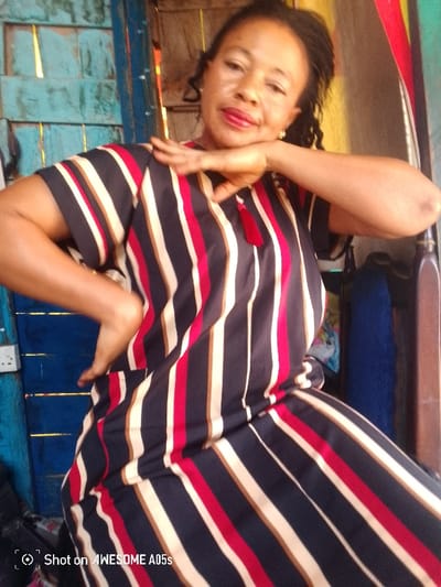 An adult female subject is depicted from the mid-torso upwards, seated or standing, in an indoor or semi-sheltered setting within Adankolo, Nigeria. The woman has dark skin, dark braided hair pulled back, and is wearing a loose-fitting garment featuring vertical stripes in navy blue, red, white, and gold/tan. The garment has a V-neckline with a red tassel detail. Her left hand is raised, with fingers splayed, and her chin rests on her palm. Her right hand is bent at the elbow, with the forearm resting on her right hip. She looks directly at the camera with a slight smile. The background on the left shows a blue wooden wall or door with horizontal planks and a white electrical outlet with two sockets. On the right, a dark brown wooden furniture piece is visible, with hints of blue fabric and other objects beyond it. The image bottom-left includes a watermark "Shot on AWESOME A05s".