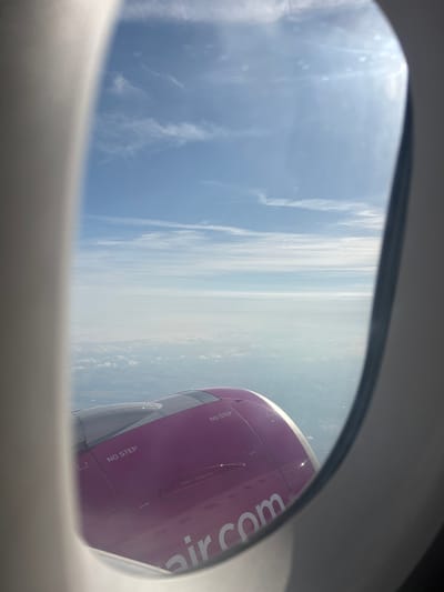 An aerial view is captured through an airplane window, showcasing the vast expanse of the sky and clouds. The lower portion of the image features the purple engine of the aircraft, with visible markings such as "NO STEP" and "NO GRAB." The text "air.com" is emblazoned on the engine. The sky above is a bright blue, dotted with wispy clouds and illuminated by sunlight, which creates a glare on the window. The environment appears to be clear and sunny, typical for an overhead flight. There is no indication of Echterdingen, Germany in the image.