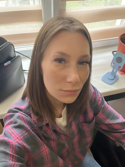 A young woman with light brown hair and blue eyes is pictured in a medium shot, looking directly at the camera. She is wearing a pink and gray plaid shirt over a white top. Her hair is shoulder-length and slightly parted. The background appears to be an indoor setting, possibly an office or a room with a window covered by blinds. To the left of the woman, a black bag is visible. To her right, a blue phone stand and a terracotta pot with a plant are on a white surface. The overall lighting suggests it might be daytime. The image does not provide any specific indicators or context that would allow for identification of the location as Taldom, Russia.