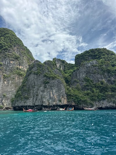 The image shows a body of turquoise water in front of a rugged, tree-covered limestone cliff face. Several longtail boats are moored in the shallow water near the base of the cliffs. The sky above is partly cloudy with a mix of white and gray clouds. The scene appears to be a scenic coastal area, likely in Thailand, given the presence of longtail boats and the distinctive karst topography.