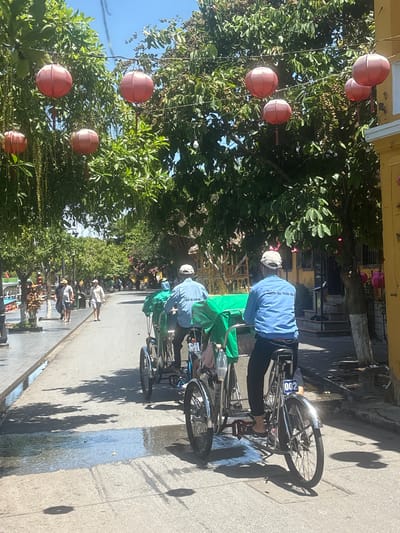 In Hội An Ward, Vietnam, on a sunny day, two cyclos are traveling down a street lined with trees and red lanterns. The cyclo in the foreground is being driven by a man in a blue long-sleeved shirt and black pants, wearing a white cap. He is pedaling the cyclo, which has a license plate reading "002". A passenger is seated in the back of the cyclo, covered by a green tarp. Another cyclo, with a driver and a passenger under a similar green tarp, is in front of the first. On the left side of the street, a paved walkway is visible with people walking and standing. The street itself appears wet in places, reflecting the dappled sunlight filtering through the trees. The architecture of the buildings on the right side of the street suggests a traditional Vietnamese style, with yellow walls and dark shutters.