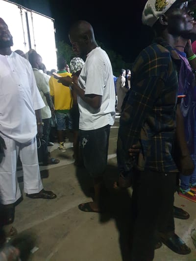 This is a nighttime outdoor scene, possibly at an event, in Lokoja, Nigeria. Several individuals are visible, some facing the camera, others with their backs turned. In the foreground, a person wearing a white robe and trousers is partially visible on the left. Next to them, another person in a white t-shirt is looking down at a mobile phone they are holding. To their right, a person in a yellow shirt and denim shorts stands, and behind them, a person in a plaid shirt and dark trousers is seen from the side. In the background, a large white truck or trailer is parked. The ground appears to be concrete or asphalt, and the lighting suggests artificial illumination from the event. The atmosphere is casual, with people gathered and interacting.