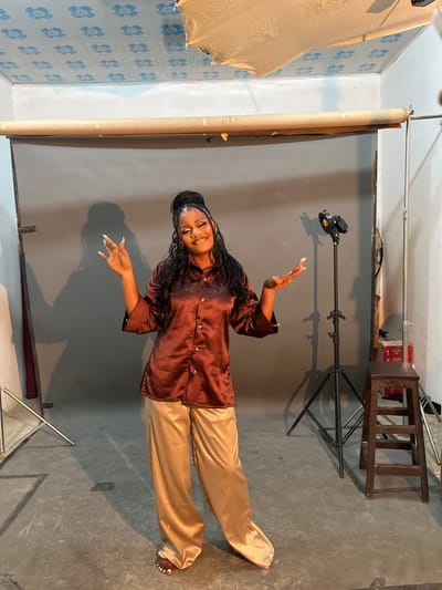 A young Black woman stands in a photography studio. She is wearing a long-sleeved, brown, satin button-up shirt and wide-legged, gold satin pants. Her hair is styled in a high bun with braids. She has dramatic makeup on, including bold eyeshadow and lip gloss. Her nails are long and painted white. Her arms are outstretched, and her hands are open, as if she is in the middle of a dance move or posing for a photo. The studio has a grey backdrop, with a roll of paper hanging from a bar behind the woman. To the right of the woman, there is a photography light on a stand, and a wooden stool. The ceiling has blue patterned tiles, and there are some lighting fixtures visible. The floor is concrete. The image does not provide specific location details within Gura Top, Nigeria, beyond it being a studio setting.