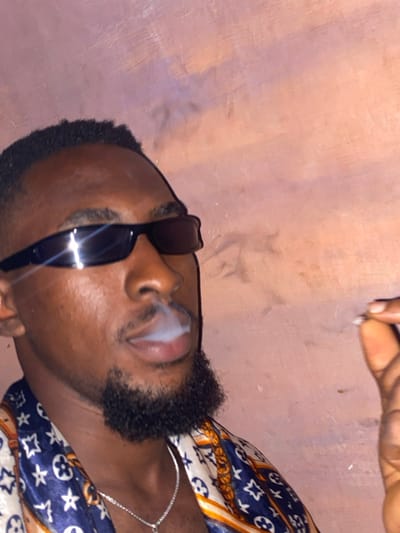 A man wearing sunglasses exhales smoke. He has dark skin, short black hair, and a styled beard. He wears a patterned shirt and a silver chain. A hand in the upper part of the image holds something small, possibly related to the smoke. The background is a textured wall, seemingly illuminated with warm light.