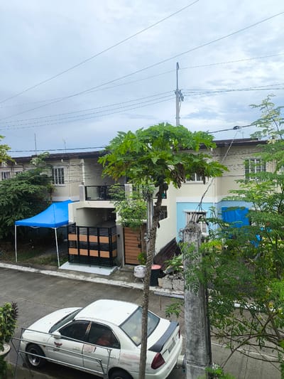 The image depicts a residential street in General Trias, Philippines, under a cloudy sky. In the foreground, a white four-door sedan is parked on the concrete road. The car exhibits some wear and tear, with visible decals on the driver's side door and a black spoiler on the trunk. Adjacent to the car is a concrete pole and lush green foliage.

Across the street, a row of houses is visible. The house closest to the viewer has a light-colored facade with brown trim and multiple windows. In front of this house, a blue tent is set up, supported by a white frame. Next to the tent, a gate with black and brown wooden panels guards an entrance. Further down the street, another house is partially visible, featuring a light yellow and blue exterior with windows and electrical wires running overhead. A papaya tree with green leaves and developing fruit stands prominently in front of the houses. The environment is characterized by concrete roads, houses, vegetation, and overhead utility lines, typical of a suburban neighborhood.
