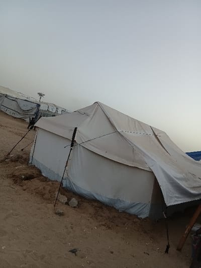 The image captures an outdoor scene featuring several large, light-colored tents under an overcast sky. The primary focus is on a prominent tent in the mid-ground, positioned slightly to the right of the frame. This tent is constructed from a white or very light grey, heavy fabric or tarp material, suggesting a durable, temporary shelter. It has a pitched roof design, with visible black ropes or guy lines extending from its sides to anchor it to the sandy ground. One such rope stretches diagonally from the tent's mid-section towards the lower left of the image. The lower part of the tent appears to be secured or reinforced, possibly by piling sand or dirt against its base, and a bluish tarp-like material can be seen layered along its top edge, suggesting a multi-layered wall or repair.
The ground surrounding the tents is predominantly dry, light brown, sandy earth, with scattered small rocks and debris, indicating a possibly arid or semi-arid environment. To the far right, partially cut off, a section of what appears to be rough wooden beams or supports is visible, hinting at other nearby structures or collected materials.
In the background, further to the left, another similar tent is partially visible, also light-colored and of similar construction, though its full extent is obscured. A small, light-colored object, possibly a small plant or a piece of debris, can be seen clinging to the side of this background tent.
The sky occupies a significant portion of the upper half of the image, presenting a uniform, pale grey-blue color, characteristic of an overcast day or perhaps twilight hours (early morning or late afternoon). There are no distinct clouds, only a flat, diffused light.
The image is captured with a tilted perspective, causing the horizon line to appear diagonally across the frame. There are no people visible, and no specific activity is taking place, although the presence of multiple tents strongly suggests a temporary settlement or camp. No discernible text is visible anywhere in the image.