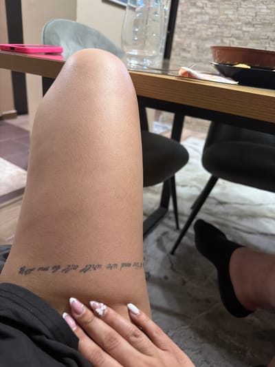 The image shows a close-up of a person's leg, with a tattoo visible on the outer thigh. The tattoo is a vertical line of text in a script font. Part of the text reads "ithe end we will all as one." The person's hand with manicured nails, featuring white tips and a small white bow design on one nail, is also visible in the frame. The background suggests an indoor setting, possibly a home or cafe, with furniture and a textured floor. A water bottle and part of a wooden table are visible on the left. The overall lighting suggests daytime.