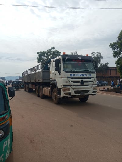 A long, white semi-truck with a trailer is parked on a paved road. The truck has "MALCOMINES LTD." written on the side of the cabin. There are also orange hazard lights on top of the cabin, and a number "80" is visible in multiple places on the truck. The trailer is empty and has a metal railing around the top.
In the background, several other vehicles are visible, including a green and yellow auto-rickshaw in the foreground on the left, and a dark green auto-rickshaw further down the road. There are also people walking and standing on the side of the road. A blue sedan is parked on the right side of the road, near a building that appears to be a shop or stall. The sky is overcast with clouds. The scene appears to be a street in a town or city, likely during daylight hours. The overall impression is one of a busy street with commercial activity. There are trees and buildings visible along the sides of the road.