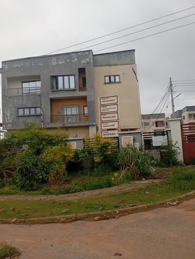 The image depicts a building under construction or renovation in Dutsen Alhaji, Nigeria. The building is a multi-story structure with a mix of grey and light yellow exterior paint, some of which appears to be peeling. The architecture features modern elements like large windows and balconies with railings. A prominent sign on the building lists project details, including "PROJECT TITLE: PROPOSED PRIVATE HOUSING ESTATE," "LOCATION: PLOT 781, B00-GHAR DISTRICT, DUTSEN-ALHAJI, GWARINPA, FCT," "CLIENT: PONGLOMERAPE LIMITED," "DEVELOPER: PARADISE ESTATE LIMITED," and "CONSULTANT: PARADISE ESTATE LIMITED." The surrounding environment includes lush green vegetation, a paved walkway, and a dirt road. Electrical wires and poles are visible, suggesting ongoing utility connections. The sky is overcast, indicating a cloudy day. No people are visible in the frame.