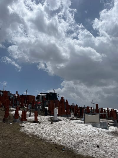 The image shows a view of a section of Noratus Cemetery, located near the village of Hartagyugh in Armenia. The scene includes numerous khachkars (Armenian cross-stones) lined up in rows on uneven ground. Some khachkars are red-orange, while others are gray or white. A portion of the ground is covered in snow. A building is visible in the background with a green roof, and scaffolding or some form of support structure is located on top of it. The sky is predominantly covered with large, gray and white clouds. There are patches of blue sky visible between the clouds.