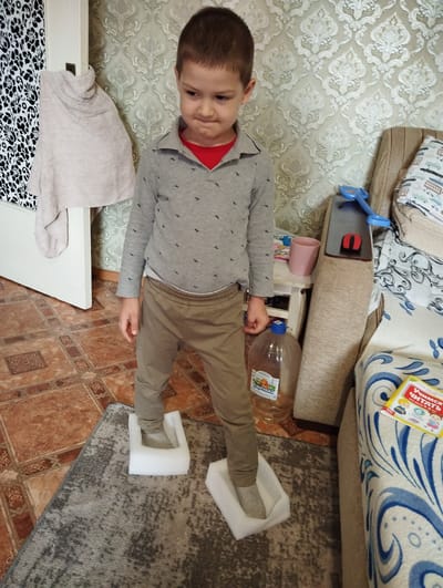 A young boy stands on two white foam blocks on a grey rug in a room. He is wearing a grey long-sleeved shirt with small dark patterns, a red collar, and khaki pants. His feet are in light grey socks. The room features patterned wallpaper and a tiled floor. To the left of the boy, a white door with a black and white patterned fabric hanging on it is visible. To the right, a beige sofa with a patterned blanket and a yellow book with Russian text are present. A large plastic bottle of liquid and a pink cup are on a small table behind the boy. The scene appears to be indoors, likely a residential setting. The city of Kingisepp, Russia is not discernible from the provided image.
