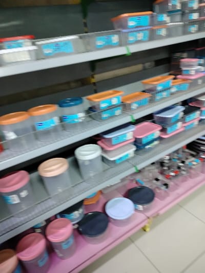 Shelves filled with plastic containers of various shapes and sizes are displayed in a store. The containers have different colored lids, including orange, pink, blue, and black. Some containers are stacked on top of each other, and there are transparent containers on the shelves. The shelving is a gray color. The bottom shelf is painted pink.