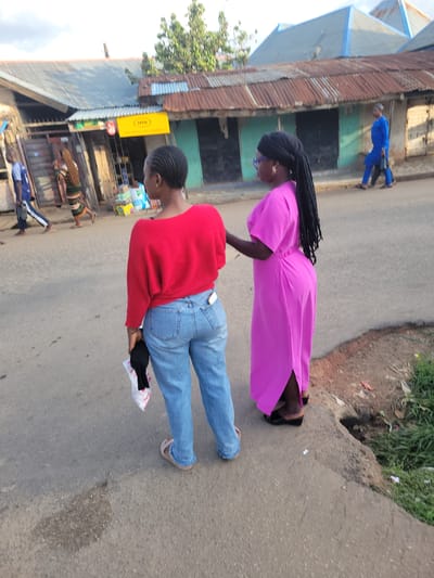 The scene depicts a street in Jos, Nigeria, during daylight hours, observed from a slightly elevated perspective behind two women in the foreground. The street surface is asphalt, with varying textures and patches.
In the immediate foreground, two individuals, identified as women, are oriented with their backs mostly towards the camera. The woman on the left wears a long-sleeved red top and light blue denim jeans with a light-colored sandal visible on her left foot. Her hair is styled in short, dark cornrows. She holds a white plastic bag containing a black item in her right hand. To her right stands another woman, dressed in a magenta-pink ankle-length dress with short sleeves and black wedge sandals. She has a dark headwrap, from which long, dark dreadlocks or braids cascade down her back. Her right arm is slightly bent at the elbow, and her hand is near the other woman's left arm. She appears to be wearing glasses with a purple frame and gold-colored hoop earrings.
In the midground and background, several other individuals are walking. On the far left, a man in a blue and white long-sleeved shirt and dark trousers walks away from the camera, carrying a dark bag. Further back and to the right of him, a woman with long braided hair is visible, wearing a vertically striped dress. On the far right, a man in a full blue traditional outfit, consistent with Nigerian attire, walks along the street.
The left side of the street is lined with commercial buildings, typically single-story structures with corrugated metal roofs of varying conditions; some are grey or blue, while others exhibit rust-brown hues. One building displays a prominent rectangular yellow sign with the text "MTN" at the top and additional smaller text below. Merchandise, including packaged goods and boxes, is visible outside some of these establishments. The street on the right edge transitions into a raised, unpaved verge with sparse green vegetation and exposed earth. A small, dark opening in the curb suggests a drainage point or culvert. The sky above is partially visible, appearing overcast or hazy.