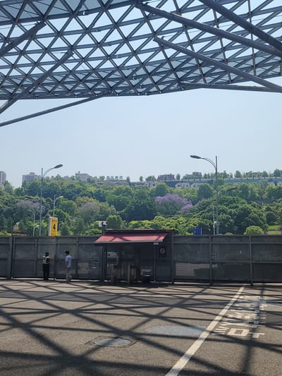 The image shows an outdoor scene in Liangjiang, China. The foreground features a complex metal structure, possibly part of a building or bridge. Adjacent to the structure is a bus stop with a shelter. Two people are present at the bus stop: one in a white shirt and light pants and another in a darker outfit, possibly security personnel. Beyond the bus stop, there is a dense area of trees with some visible buildings in the background. The road surface in the foreground has Chinese characters painted on it and is covered in the shadows cast by the metal structure. The sky is clear and blue.