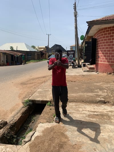 The image captures a full-body shot of a young man standing on a concrete roadside in what appears to be an urban or semi-urban setting in Jos, Nigeria, under bright daylight. The man, wearing a red short-sleeved t-shirt, dark trousers, and dark sandals, stands with his hands clasped together near his chest, looking towards the camera. He casts a long, distinct shadow on the dusty ground beside him, suggesting either early morning or late afternoon sunlight.
To his right (viewer's left), a paved road stretches into the distance, with what look like market stalls or small shops along its edge. To his immediate left (viewer's right), a concrete drainage ditch runs parallel to the road. This ditch contains some water, dirt, and scattered green vegetation.
In the background, a two-story building with a red brick facade and a corrugated metal roof is visible. Below this building, there's a power pole laden with numerous electrical wires and utility boxes, along with a green satellite dish attached to a lower part of the building. Several signboards are visible, one of which clearly displays the text "THE GREAT". A motorcycle is parked near the pole. Further in the background, other buildings with varying architectural styles, including a structure with a white corrugated roof, are visible under a clear, pale blue sky.
The scene depicts a typical street environment in the region, characterized by its infrastructure, building materials, and the strong tropical sunlight. The man is the central figure, actively posing for the photograph.