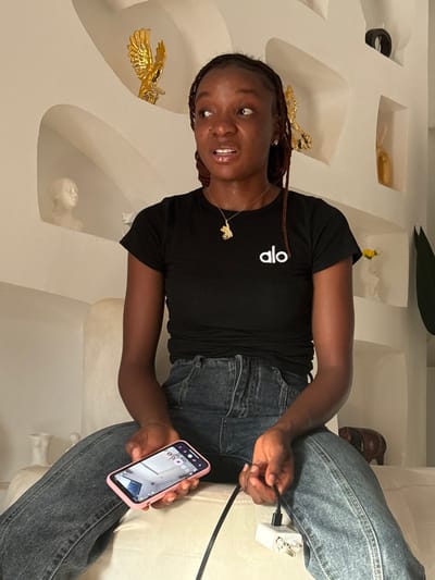 An individual is seated indoors in Lafia, Nigeria, on a light-colored upholstered surface against a decorative white wall featuring multiple recessed niches. The wall niches display various ornamental objects, including a golden eagle statuette, a white bust sculpture, a golden abstract sculpture, a small yellow flower in a white vase, and a dark brown animal figurine. The seated individual is a female with dark skin and braided dark hair, wearing a black short-sleeved t-shirt with a white "alo" logo on the left chest and blue denim jeans. She wears a gold-colored pendant necklace. Her mouth is slightly open, and her eyes are wide, directed towards the upper right of the frame. In her left hand, she holds a pink-cased smartphone with its screen illuminated, displaying an interior room. In her right hand, she holds a black charging cable connected to a white two-pronged wall adapter.
