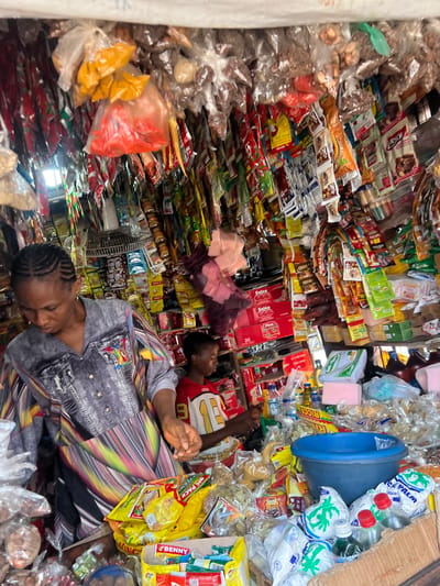 The image depicts a street market stall in Nkpor, Nigeria, overflowing with a wide variety of goods. The stall is densely packed with products ranging from food items, such as spices in plastic bags and packaged snacks, to bottles of liquids and what appear to be detergents. The items are hung from the ceiling and stacked on shelves, creating a visually busy environment. Two individuals are visible: a woman in the foreground, wearing a grey and colorful patterned shirt, appears to be attending to the stall, and a younger person is in the background wearing a red and white shirt. The lighting suggests it is daytime, with natural light filtering into the stall. The overall impression is one of a vibrant and active marketplace.