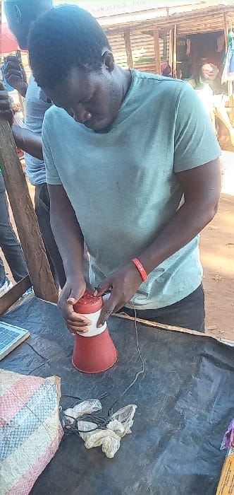 A young man is standing in front of a wooden structure, wearing a light blue t-shirt and a red bracelet on his left wrist. He is looking down at a red and white object in his hands, likely a speaker. There is a black cable connected to the speaker and a solar panel on the table. The man is holding a speaker in front of a wooden structure, possibly a shop or a home. The scene takes place in Lwandeti, Kenya, a rural area characterized by simple structures and natural surroundings. The man is likely setting up the speaker for some purpose, possibly for music or announcements. The scene suggests an informal setting in a rural part of Lwandeti, Kenya.