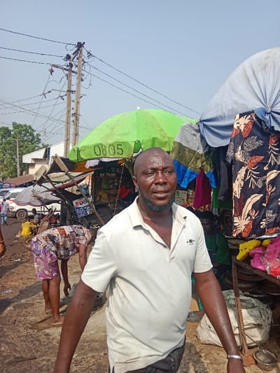 This image depicts a bustling outdoor market scene in Onitsha, Nigeria, under clear daylight.
In the immediate foreground, a dark-skinned adult male with a bald head and a short beard is positioned slightly off-center, facing towards the viewer with a neutral expression. He wears a light-beige short-sleeved polo shirt and dark shorts. Sweat is visible on his forehead, face, and arms. A simple silver-colored bracelet adorns his left wrist.
Behind this individual, the scene transitions into a dynamic market environment. To the left, another individual, wearing distinctive patterned shorts (purple, white, brown, and black) and barefoot, is bent over, engaged in an activity on the unpaved, uneven ground. Further left, a white van or bus is partially visible amidst other indistinct figures and market structures. A man in a yellow shirt is seated behind a small market cart.
To the right of the foreground figure, several market stalls are present. One prominent stall features a bright green umbrella with the number "0805" inscribed in white. Adjacent to it, another stall is covered by a light blue/grey umbrella. Clothing items, including a dark shirt with a light-colored leaf pattern, are displayed hanging from these structures. On a lower surface to the right, various items, notably bright yellow and pink molded plastic objects (possibly shoes or toys), are arranged for sale. A large, dirty white sack rests on the ground nearby. A partially obscured figure in a green shirt is visible behind the rightmost stall.
Overhead, multiple utility poles with a dense network of electrical wires crisscross the bright, clear sky. In the background on the far left, green foliage and trees are visible, along with parts of low-rise buildings. The ground throughout the market area appears to be unpaved, characterized by dirt and debris.