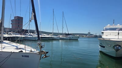 The image depicts a harbor scene on a clear, sunny day. In the foreground, the bow of a white sailboat with the registration "Vn 8666" is visible. Behind it, several other sailboats and yachts are moored, with tall masts reaching towards the blue sky. A modern, multi-story building with red and grey accents stands on the shore in the mid-ground, likely part of the Varna Sea Station or a related facility. Further in the background, more vessels, including what appear to be naval ships and a lighthouse, are visible along the coastline. The water is calm and reflects the sky and surrounding boats. The scene is consistent with the port of Varna, Bulgaria, given the architecture and the presence of a mix of recreational and potentially commercial or naval vessels. No people are clearly visible in the image.