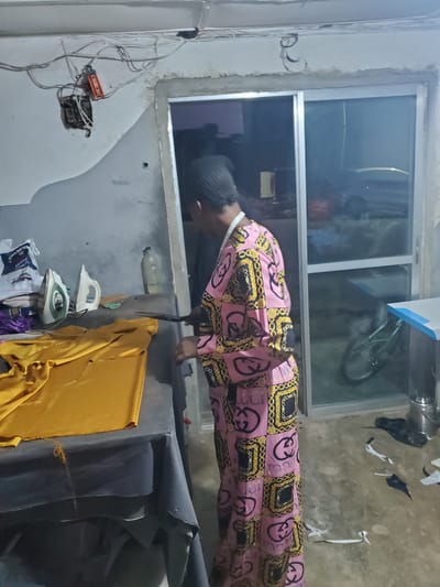 A person wearing a pink and gold patterned kaftan is leaning over a table, seemingly engaged in some activity. The person has dark skin and is wearing a dark cap. A measuring tape is wrapped around their neck, suggesting they might be a tailor or seamstress. The setting appears to be a workshop or a domestic space with construction elements. In the background, a window shows a reflection of a bicycle.
On the table, there are two irons, one white and one green and white, placed on top of a yellow fabric. A clear plastic bottle is also visible on the table. Various wires and an electrical junction box are mounted on the wall to the left, indicating ongoing or recent electrical work. The floor is rough concrete. The overall scene suggests a somewhat cluttered and utilitarian environment, possibly a tailoring shop in Jos, Nigeria. The lighting suggests it is daytime, but the interior lighting is the primary source. No weather cues are apparent. There is no visible text in the image.
