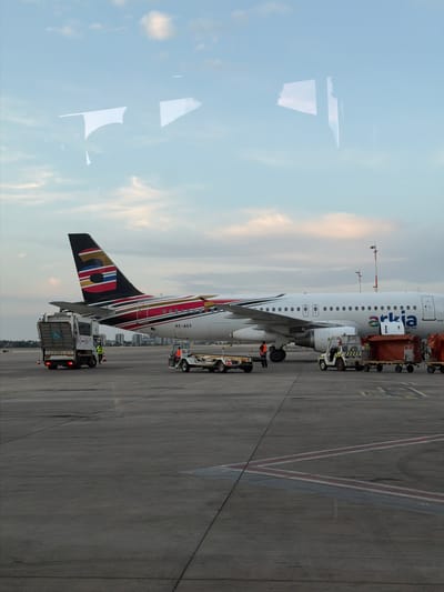 An airplane is parked on an airport tarmac, viewed through a window, evident by prominent reflections of the sky and clouds on the left side of the image. The aircraft, a white narrow-body jet, features "Arkia" written in both Latin and Hebrew script on its fuselage. Its tail fin is distinctive, adorned with a colorful design incorporating red, black, white, gold, blue, and green stripes and patterns. The registration number "4X-AGY" is visible near the tail section.
Around the aircraft, several ground support vehicles are present, indicating active airport operations. A large service vehicle, possibly a catering or cleaning truck with "RENO LIFT" visible on its side, is positioned near the aircraft's tail. Other ground vehicles, resembling baggage tugs or loaders, are seen closer to the main fuselage and nose area. Ground personnel are also visible, engaged in tasks around the aircraft and vehicles.
The tarmac is made of concrete, marked with various painted lines in white, red, blue, and yellow, typical of airport aprons. In the background, beyond the aircraft, a distant urban skyline or airport buildings are faintly visible under a soft, overcast sky, suggesting either early morning, late afternoon, or a heavily clouded day. The overall scene depicts typical ground operations at an airport in Yagel, Israel.