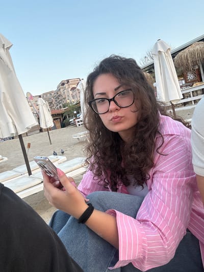 A young woman with curly brown hair and black-rimmed glasses is captured in a close-up selfie on a beach. She is wearing a pink and white striped shirt and blue jeans. Her mouth is puckered as if she is about to kiss. She is holding a smartphone in her left hand. The background shows a sandy beach with white lounge chairs and umbrellas, with buildings visible in the distance. This scene is located in Sveti Vlas, Bulgaria.