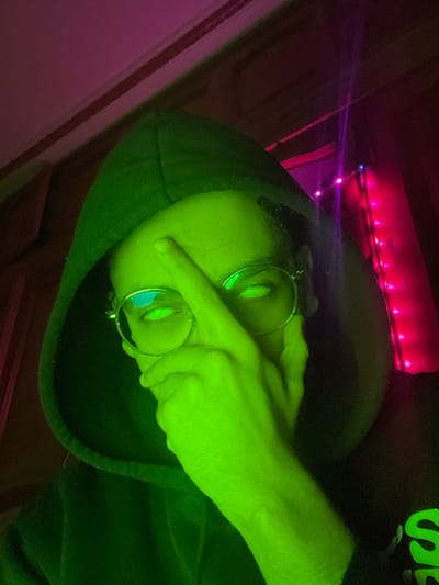 This is an indoor, close-up, chest-up portrait of an individual located in the city of Fez, Morocco, captured under artificial and stylized lighting conditions. The subject wears a dark hooded sweatshirt and round, wire-rimmed glasses, with their face and visible hand strongly illuminated by an intense neon green light source. The eyes, seen through the glasses, reflect or emit a bright green luminescence. The individual's right hand is raised, with the index finger touching the center of the forehead and the thumb resting against the jawline. A fragment of a black garment featuring neon green lettering is visible on the subject's lower right side, beneath the hoodie. The background is dark and indistinct, with vertically arranged pink and purple string lights visible on the right side of the frame.