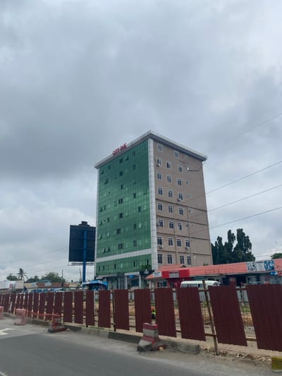 The image shows a tall building with a green glass facade and a sign that reads "CITY ONE". The building is located on the corner of a street, with a fence and several vehicles parked in front of it. There are several other buildings in the background, including a billboard with a blue background. The sky is overcast with clouds, and there are power lines running overhead. The image was taken in Dar es Salaam, Tanzania, at the coordinates -6.773, 39.241. The scene appears to be on a busy street in a commercial area, with a mix of modern and older buildings. There are no people visible in the image.