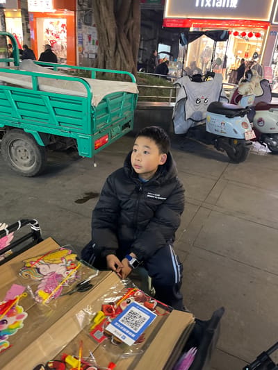 An elevated view captures an evening street scene in Jiangjin, China, with a young male child as the focal point. The child, appearing approximately 7-10 years old, sits on a low stool or cushion on a paved surface, wearing a black puffer jacket with the text "YOUTH IS BRIGHT AS SUNSHINE" and dark blue track pants with light blue stripes. A blue and white smartwatch is visible on his left wrist. He looks towards the upper right with a neutral expression, his hands clasped in front of him.

Immediately in the foreground, to the child's left, is a makeshift display or table showcasing various items within clear plastic bags, including colorful unicorn-themed lanterns or toys, and small craft items. A black pair of scissors rests on the display, along with a sticker featuring a blue QR code, likely for mobile payment.

In the midground, to the child's left, a green three-wheeled utility vehicle with a cargo bed covered by a light-colored tarp is parked. To the child's right and slightly behind him, two electric scooters are parked; the closer one is light blue with cartoon decals, a white protective handlebar cover featuring cartoon eyes, and a visible license plate reading "豫S 1515B".

The background reveals a bustling urban environment illuminated by artificial light. Storefronts line the street, including one on the center-right with "TIXIAN" prominently displayed in English and Chinese characters, containing red lanterns. Another orange-lit storefront is visible on the far left. A large tree trunk stands between the utility vehicle and the storefronts. Several blurred adult figures are visible in the background, walking or standing. The ground is paved concrete.