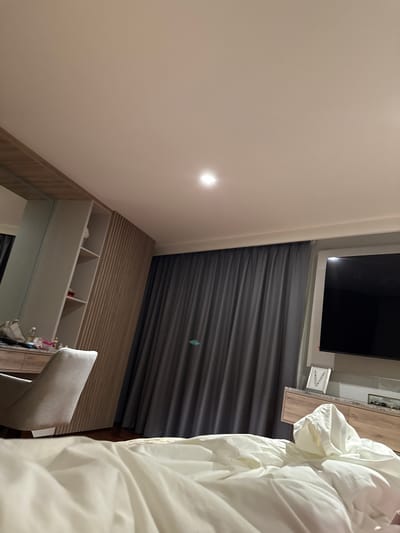 The image depicts a bedroom interior. In the foreground, a rumpled white duvet is visible, suggesting a bed. To the left, a light-colored upholstered chair is positioned in front of a wooden dresser with drawers. Behind the chair, a wall unit with shelves is present, and a large mirror covers a portion of the wall. Further back, dark gray curtains hang across a window. To the right, a large flat-screen television is mounted on the wall above a wooden cabinet. A framed picture with floral motifs is placed on the cabinet. The room is lit by a ceiling light fixture. The location is identified as San Bernardino Tlaxcalancingo, Mexico.