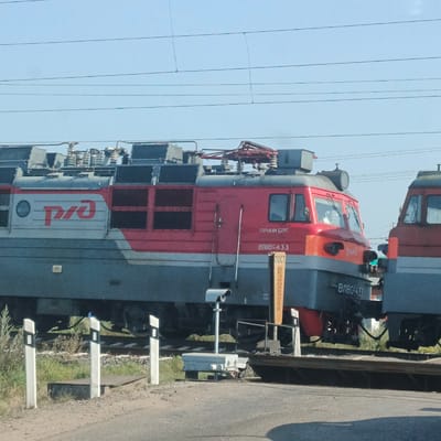 An exterior medium shot captures two red and gray electric locomotives of Russian Railways on a bright, clear day. The locomotive on the left is closer to the viewer and features the Russian Railways logo, a red circular emblem with stylized "RZD" in white. The number "ВЛ80-433" is visible on its side. The locomotive on the right is partially visible, situated to the right and slightly behind the first. Both locomotives are positioned on railway tracks, with overhead power lines visible against the sky.

In the foreground, a road crosses the railway tracks. Several white and black striped road markers, typical of level crossings, are positioned along the road. A security camera is mounted on a pole near the tracks. A yellow sign with vertical black lettering, reading "ПОСТ БЕЗОПАСНОСТИ" (POST BEZOPASNOSTI, meaning "Safety Post"), is also present. The ground is a mix of asphalt and gravel, with patches of green grass and vegetation on either side of the tracks. The scene is likely at a railway crossing in the city of Kambarka, Russia, given the context. No people are visible in the image.