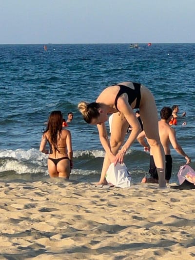 The image depicts a beach scene in Da Nang, Vietnam, with several individuals in the water and on the sand. In the foreground, on the sandy beach, a person wearing a black swimsuit is bent over, holding a white bag. To their left, another person in a black bikini stands in the shallow water, facing away from the camera. In the background, several other people are visible in the ocean, some with buoys. The water is blue with moderate waves, and the sky is a pale blue. The scene appears to be a casual day at the beach.