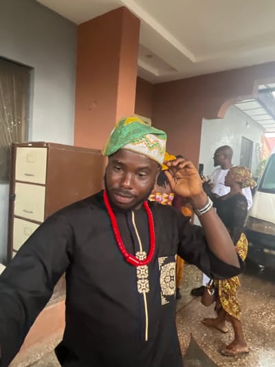 In an outdoor setting, likely a residential compound in Jos, Nigeria, a man is the central subject, dressed in traditional attire. He wears a black garment with intricate gold embroidery and a vibrant, multi-colored beaded hat. A prominent red beaded necklace adorns his neck. His right hand is raised, touching his hat. The background reveals a beige filing cabinet against a textured wall. Other individuals are partially visible, including a man in a white garment holding a phone and a woman in a patterned dress. The ground is paved with concrete. The lighting suggests an overcast day or late afternoon.