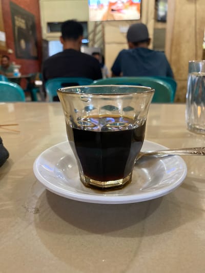 A close-up shot shows a glass of dark liquid, likely coffee, on a small white saucer. A spoon rests on the saucer. The glass and saucer are placed on a light-colored, textured table. In the background, out of focus, two men are seated, facing away from the camera. They are in what appears to be a cafe or restaurant, with blurred chairs and tables visible. A television screen with colorful images is mounted on the wall in the background. The overall ambiance suggests a casual, public dining setting. The location is identified as Lhokseumawe, Indonesia, implying this scene is taking place within the city.