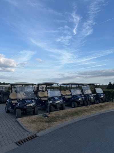 A line of dark blue golf carts is parked on a paved surface in front of a hedge and a tree line under a cloudy blue sky. The golf carts have beige roofs and seats. Some of the carts are plugged into charging stations. The ground in front of the carts is covered in dry grass. A drain is visible on the left side of the paved area. The setting appears to be a golf course or a similar recreational facility in Méan, Belgium. There are no people clearly visible in the image.
